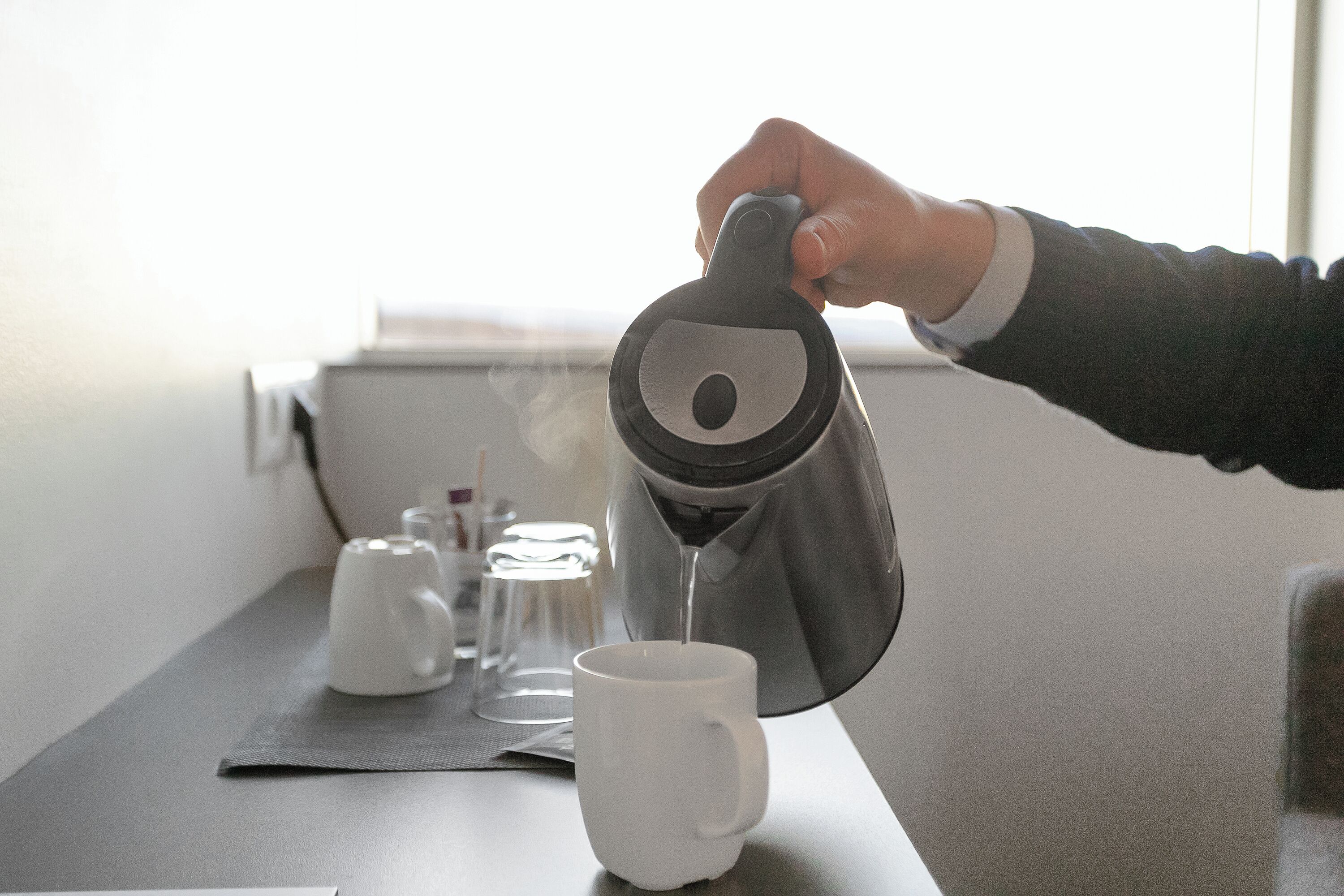 Eine Hand gießt Kaffee in eine weiße Tasse auf einem Tisch mit einer grauen Unterlage, einem weißen Teekessel und einem Glas Wasser.