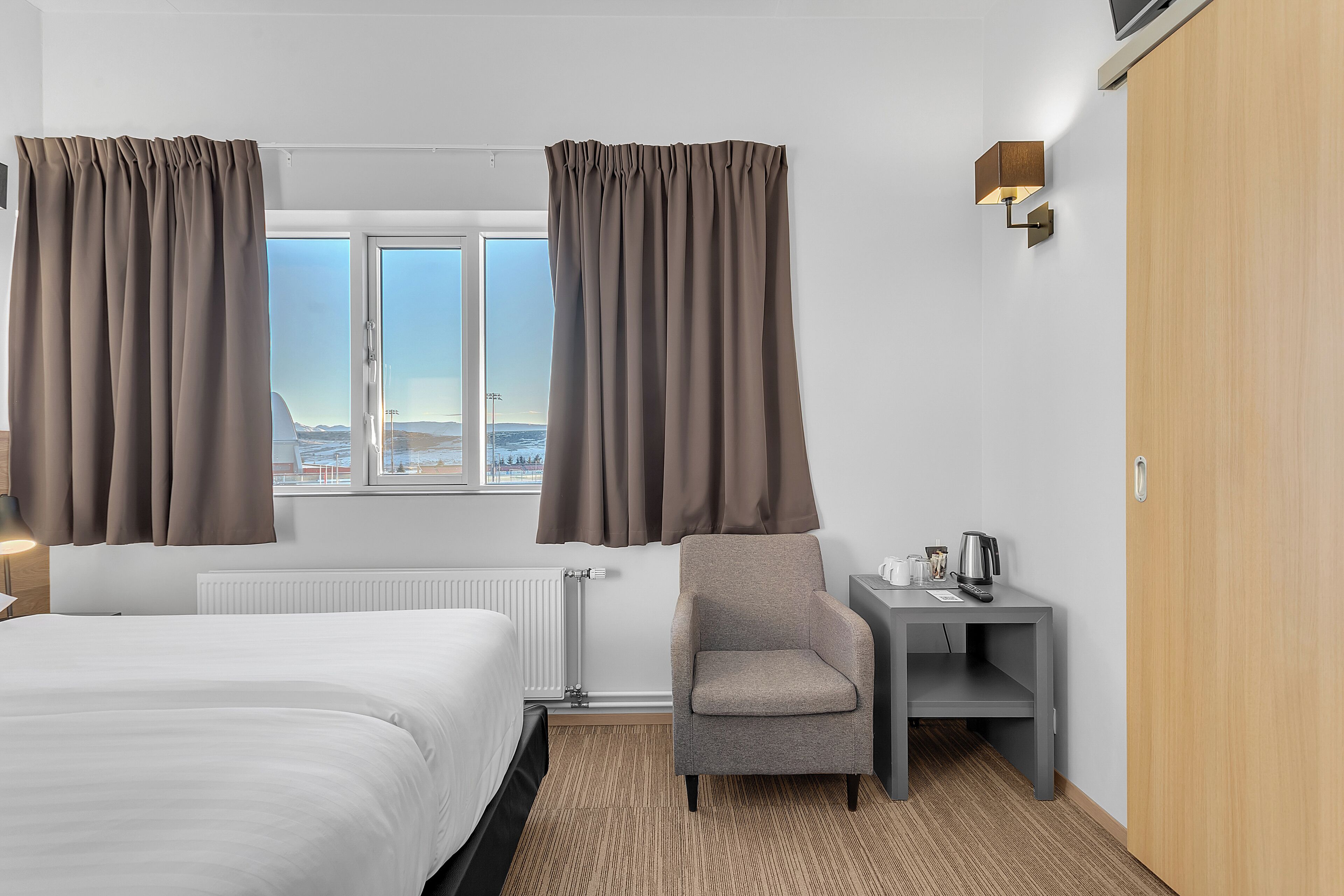 Habitación de hotel con cama, una silla, una mesita con tetera y vistas a la ciudad por la ventana.