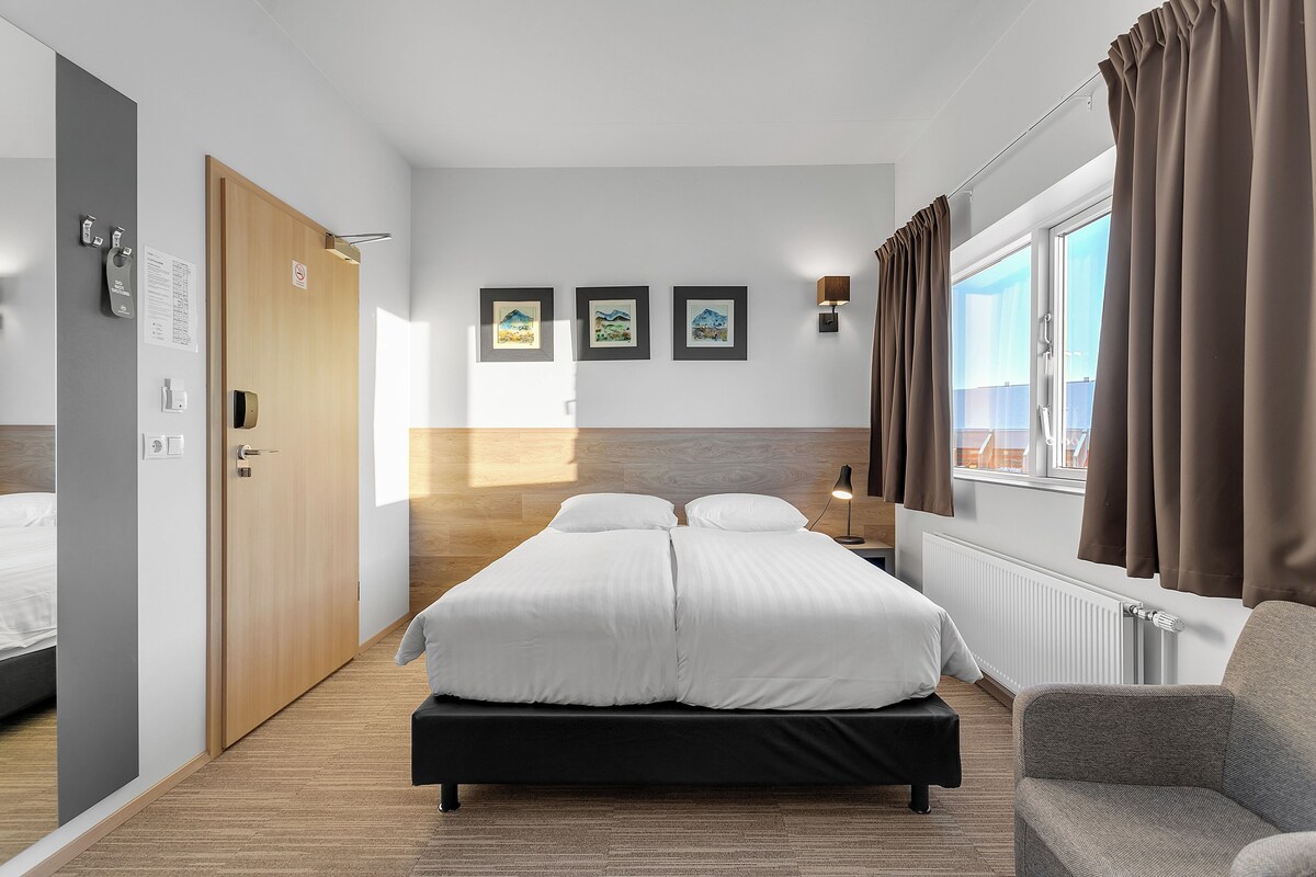 Una habitación de hotel con una cama, una silla, un ventanal con cortinas y tres cuadros en la pared.