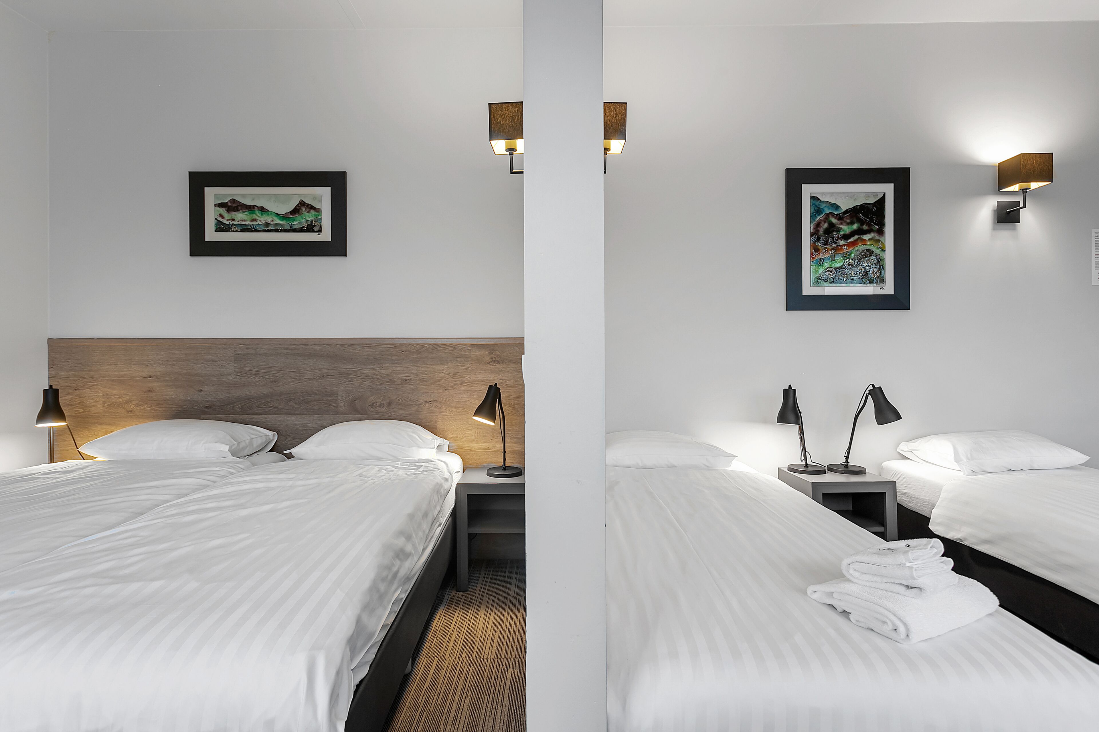 Une chambre d’hôtel avec deux lits, une tête de lit en bois, une table de chevet avec une lampe et deux lampes fixées au mur.