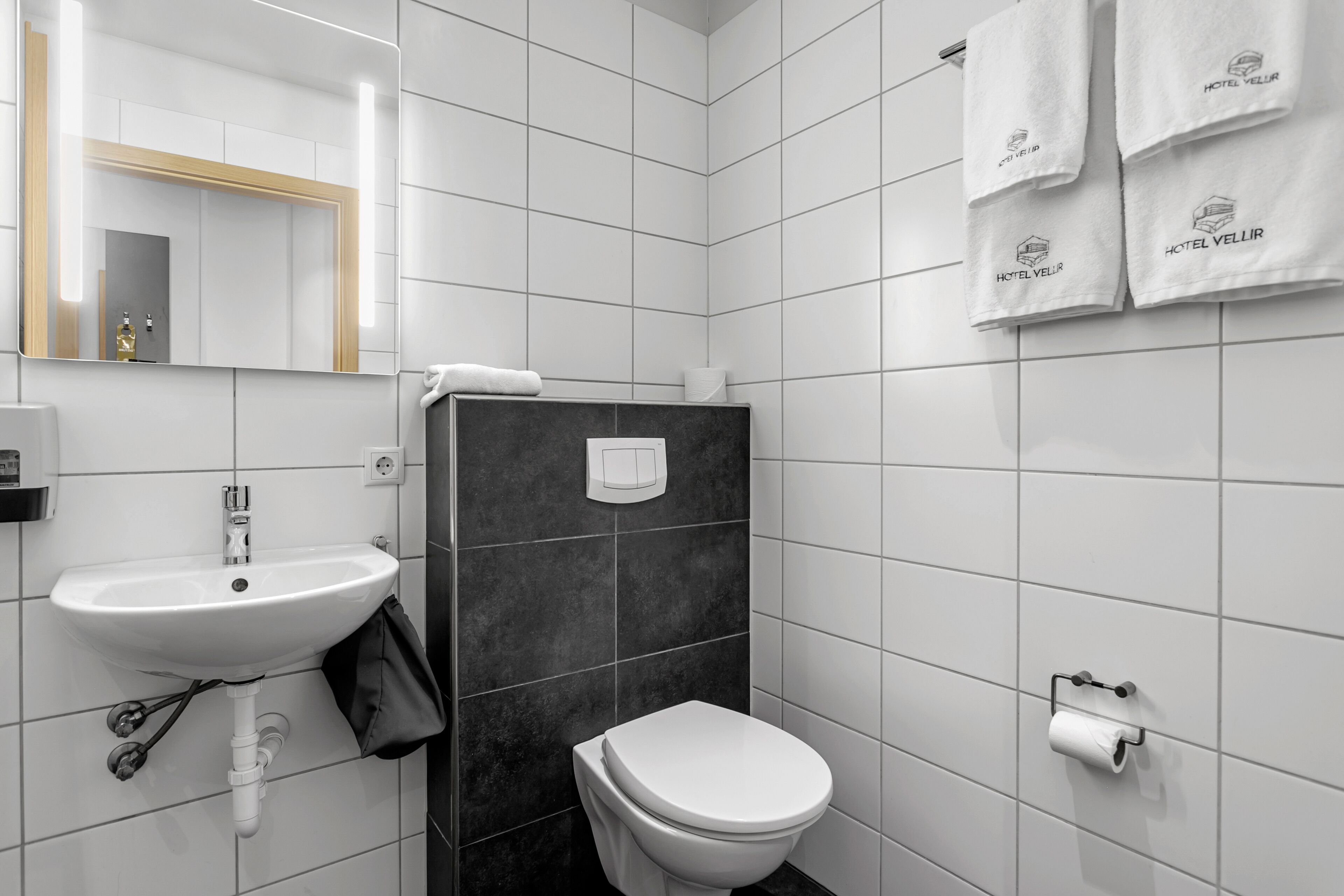 A modern bathroom with a white sink, a toilet, and a mirror. There are towels hanging on the wall and a toilet paper holder.