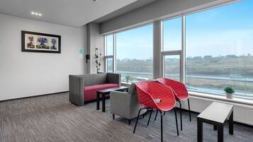 A modern office space with a gray sofa, red chairs, a small table, and a large window offering a view of a cityscape.
