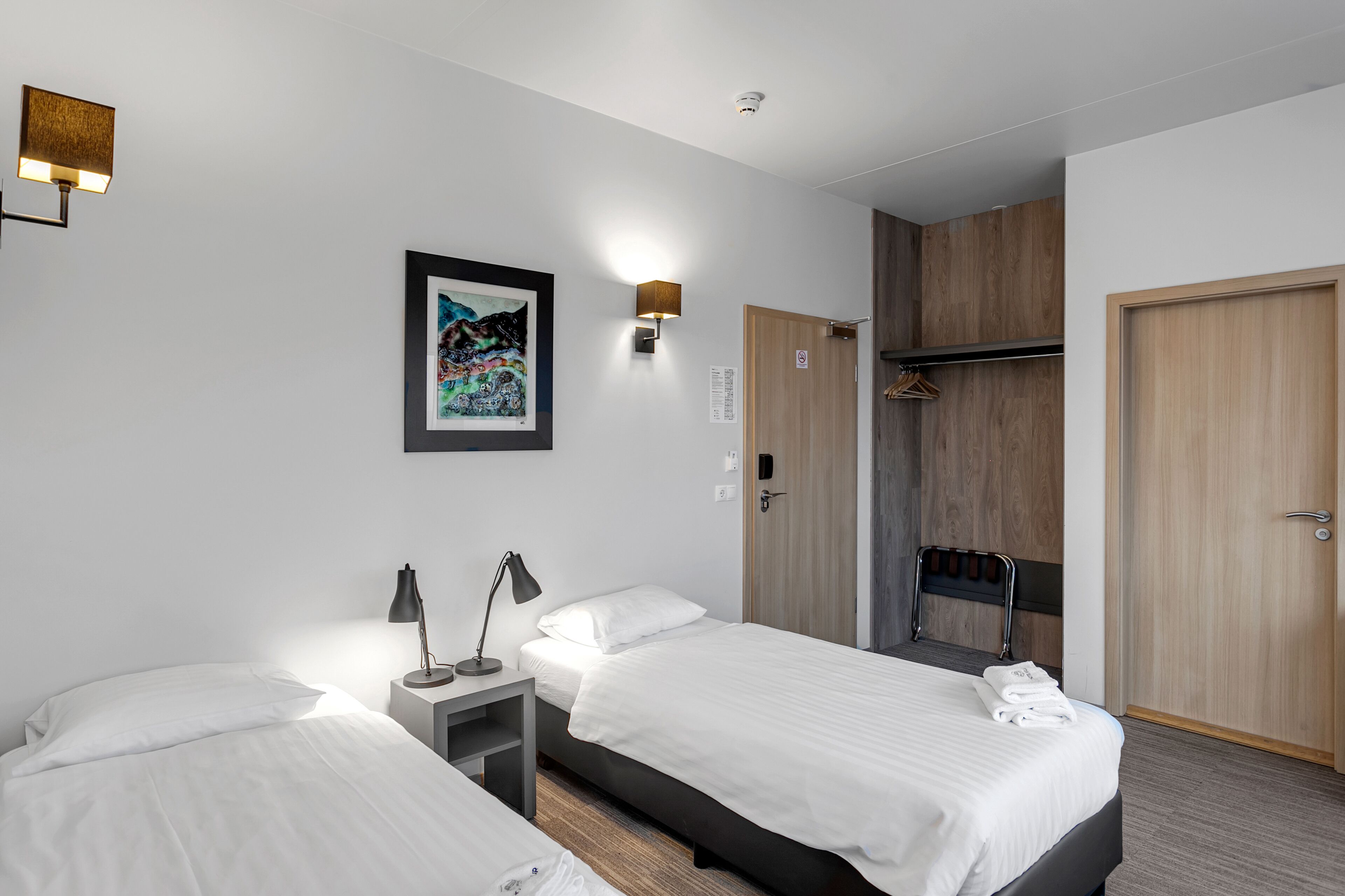 A hotel room with two beds, a bedside table with a lamp, a framed artwork on the wall, a wooden wardrobe, and a shower area.