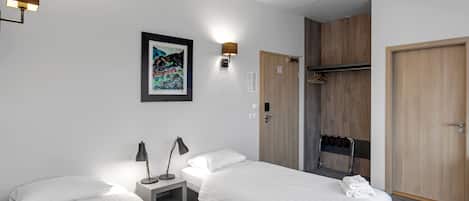 A hotel room with two beds, a bedside table with a lamp, a framed artwork on the wall, a wooden wardrobe, and a shower area.