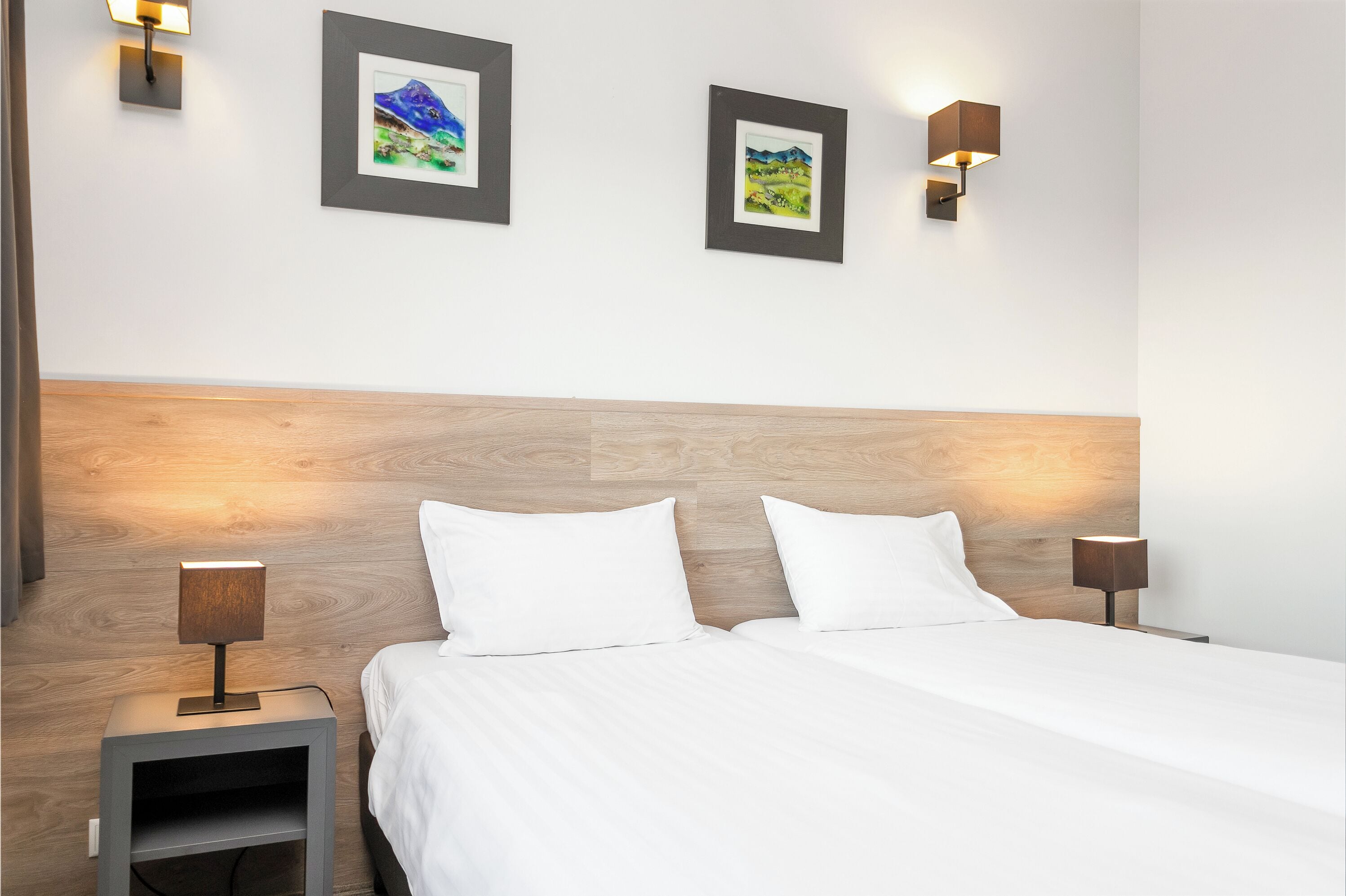 Habitación de hotel con cabecera de madera, dos mesitas de noche con lámparas y dos cuadros en la pared.