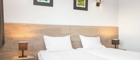 Habitación de hotel con cabecera de madera, dos mesitas de noche con lámparas y dos cuadros en la pared.