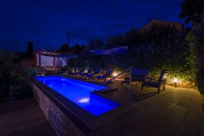 Uma piscina com iluminação azul na borda, cercada por espreguiçadeiras e um muro de pedra.