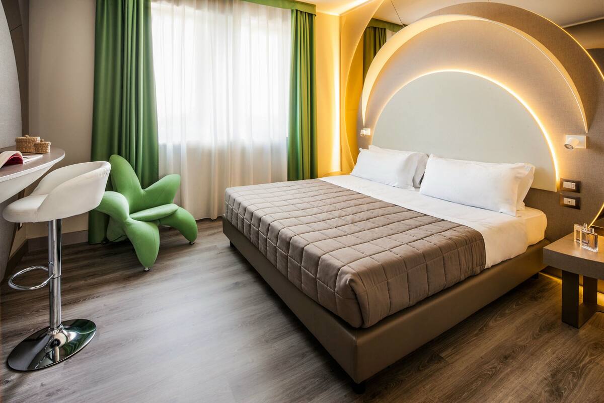 A modern hotel room with a large bed, a desk with a chair, and a green armchair.