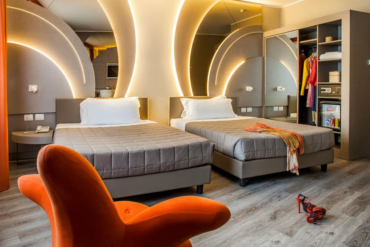 A modern hotel room with two beds, a bright orange chair, and a wardrobe with hanging clothes.