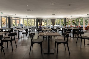 Een ruime eetruimte met meerdere tafels en stoelen, grote ramen met uitzicht op bomen en een modern plafondontwerp.