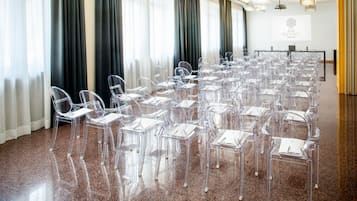 A conference room with clear plastic chairs arranged in rows, a projector screen, and a sign for Hotel Da Vinci Milano.