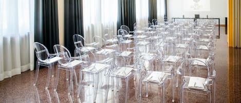 Une salle de conférence avec des chaises en plastique transparent disposées en rangées, un écran de projection et un panneau indiquant l’hôtel Da Vinci de Milan.