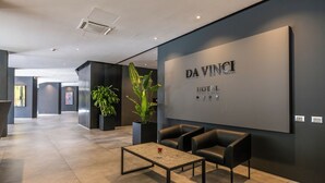 A modern lobby with a large wall sign reading "DA VINCI HOTEL," a seating area with black leather chairs, a marble coffee table, and potted plants.
