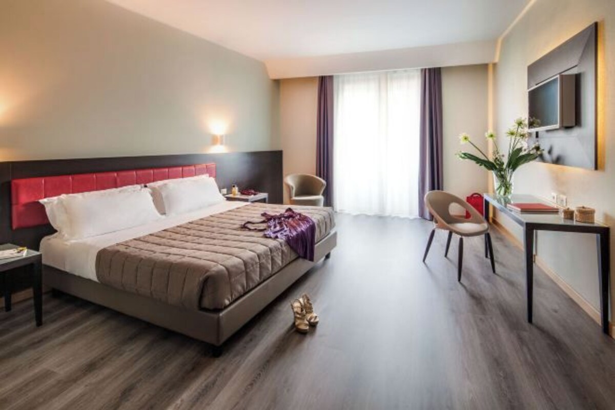 Habitación de hotel con una cama grande, un escritorio, dos sillas, un televisor y una ventana con cortinas.