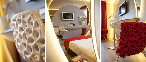 A modern hotel room with a curved bed, a flat-screen TV, a desk with a chair, and a red decorative chair.