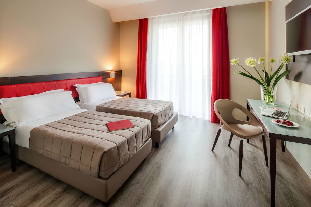 A hotel room with two beds, a desk with a laptop, a vase with flowers, and a red curtain.