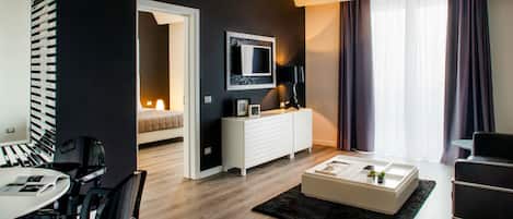 Een moderne woonkamer met een witte bank, een salontafel en een televisie aan de muur. Er is een eethoek met een tafel en stoelen, en een gang die leidt naar een slaapkamer.