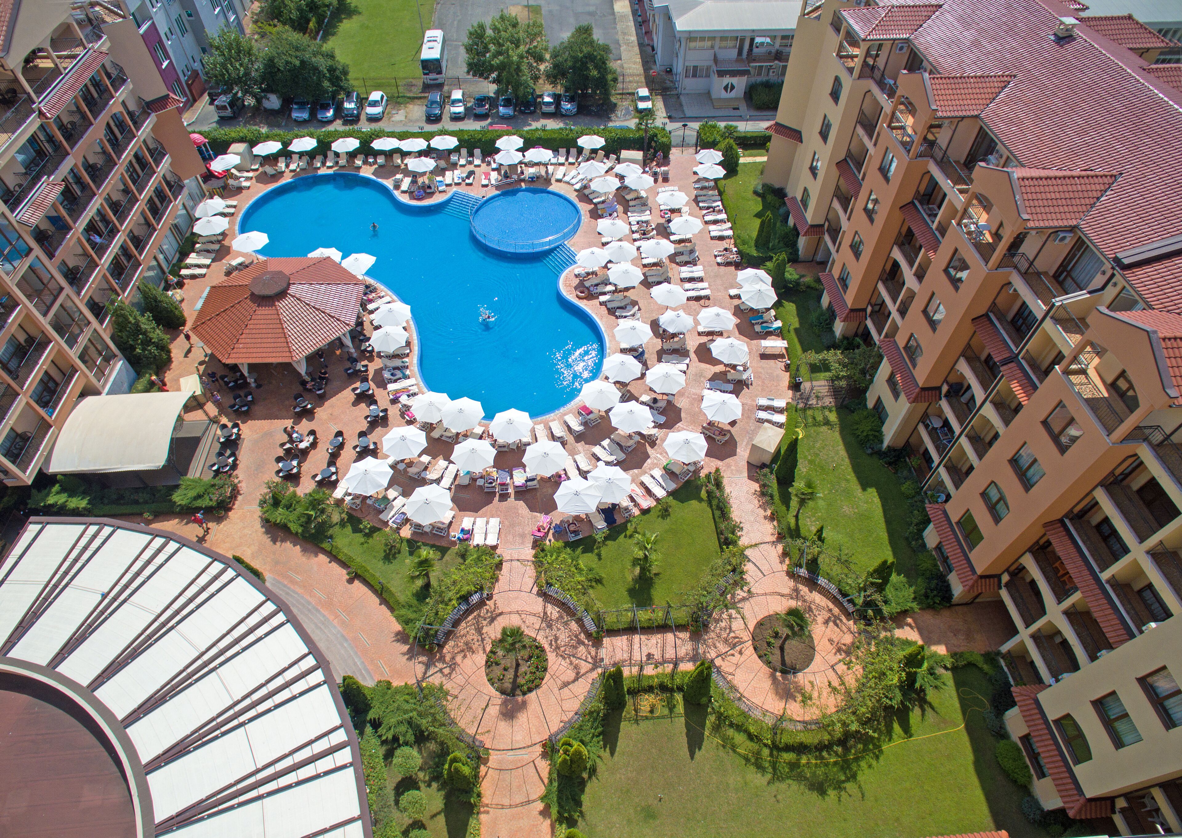 Ein Hotel mit einem zentral gelegenen Schwimmbad, umgeben von Liegestühlen und Sonnenschirmen, einem überdachten Sitzbereich und einem angelegten Garten mit einem Rundgangweg.