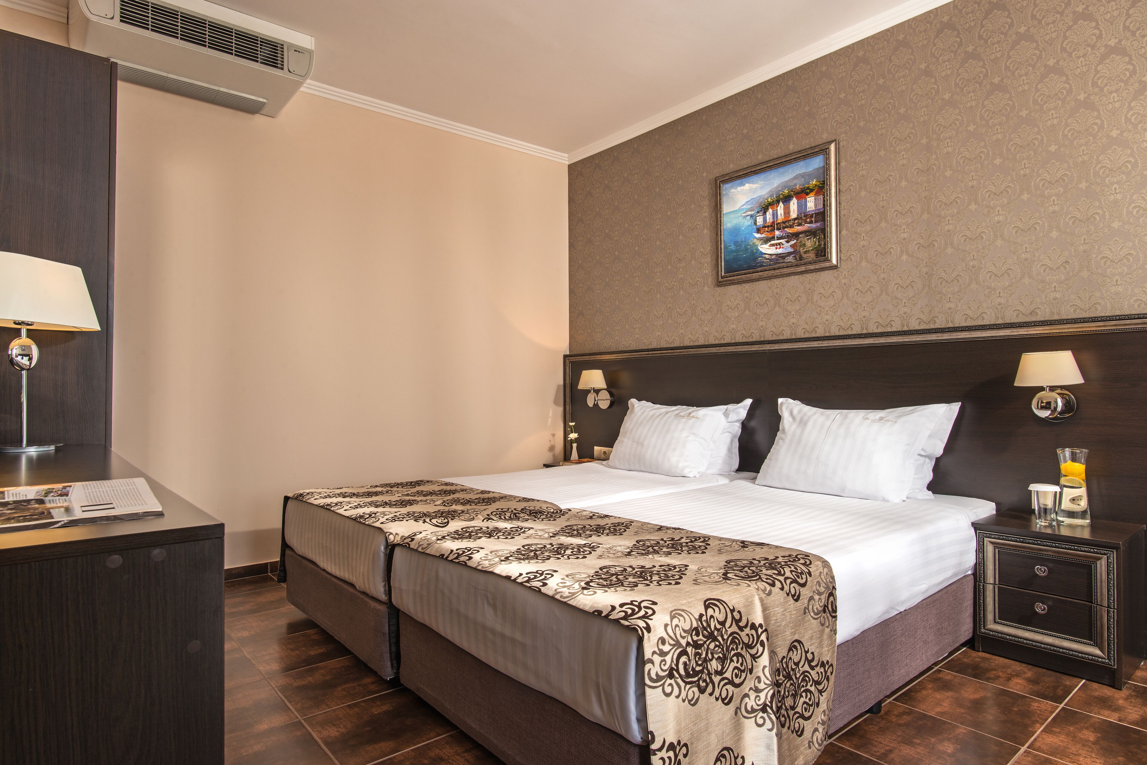 A hotel room with a large bed, a desk, a nightstand, and a wall-mounted air conditioner.