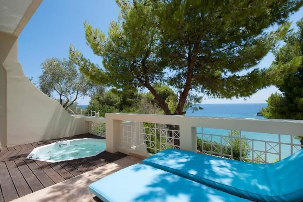 Eine Holzterrasse mit Whirlpool und einer blauen Liege, mit Blick auf eine Küstenlandschaft mit Bäumen und dem Meer.