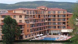 A multi-story hotel with balconies, a swimming pool, and a view of the surrounding hills. - Apart-hotel Happy (Sunny Beach)