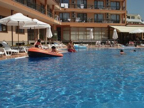 A swimming pool area with lounge chairs and umbrellas, a lifeguard on a rescue board, and a building with balconies in the background. - Apart-hotel Happy (Sunny Beach)