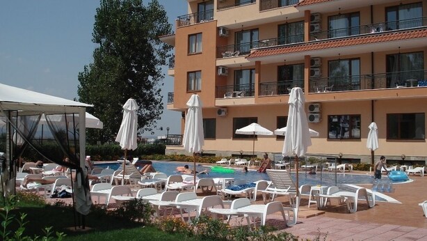 A pool area with lounge chairs and umbrellas, a covered seating area, and a multi-story building with balconies. - Apart-hotel Happy (Sunny Beach)