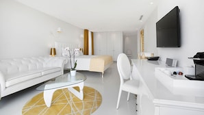 A modern hotel room with a white sofa, a glass coffee table, a bed with a yellow and white striped blanket, a white desk with a laptop, and a flat-screen TV mounted on the wall. - Portals Hills Boutique Hotel (Calvia)