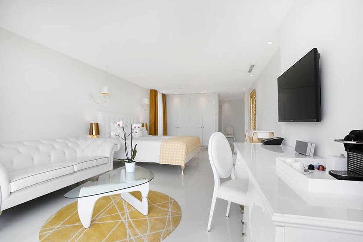 A modern hotel room with a white sofa, a glass coffee table, a bed with a yellow and white striped blanket, a white desk with a laptop, and a flat-screen TV mounted on the wall.