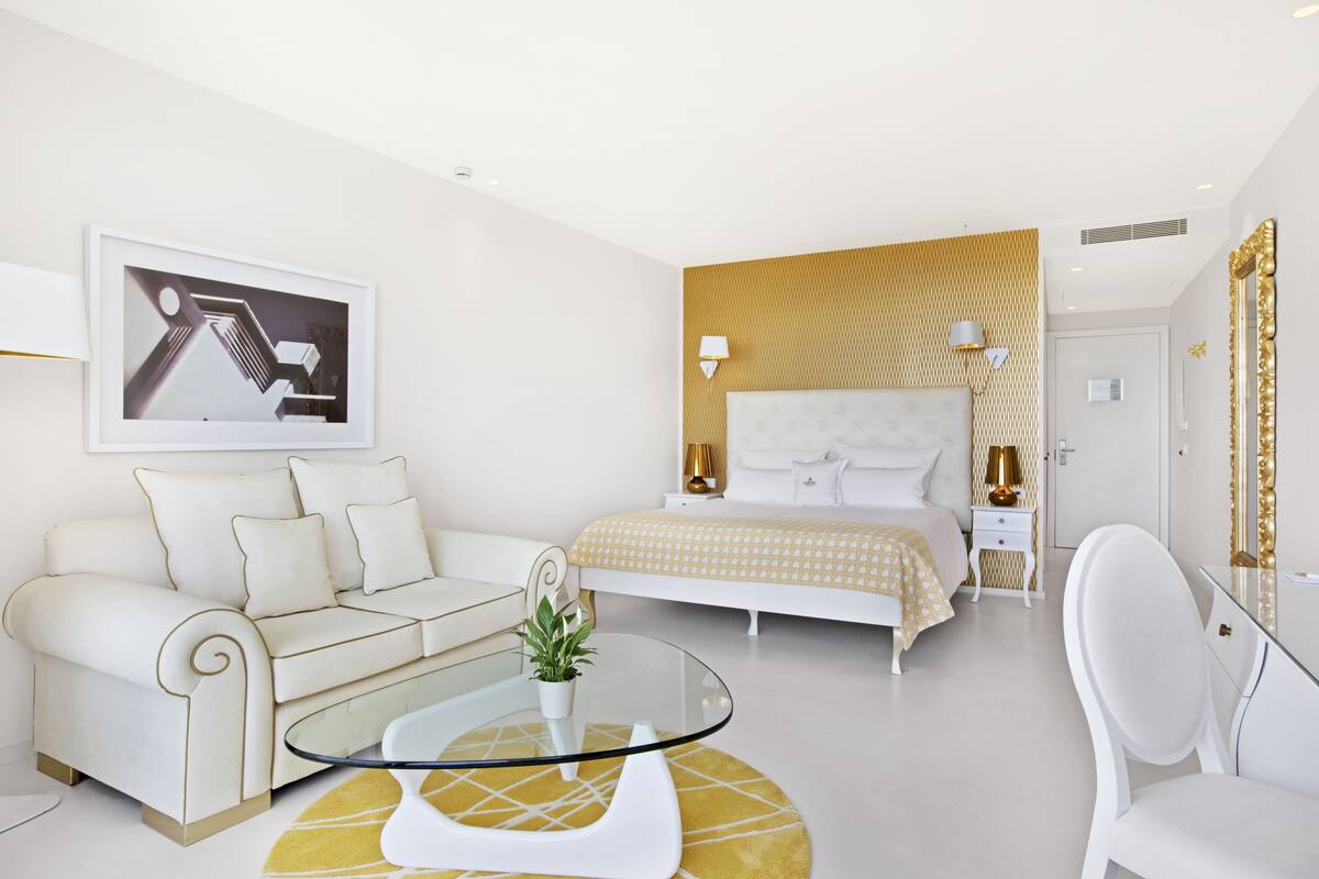 A modern hotel room with a white sofa, a glass coffee table, a bed with a patterned headboard, and a large mirror with a gold frame.