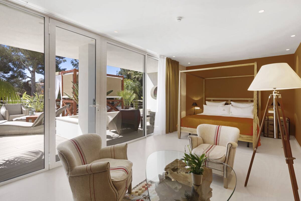 A modern hotel room with a glass door leading to a balcony, a bed with white linens, a standing lamp, and a glass coffee table with a plant.