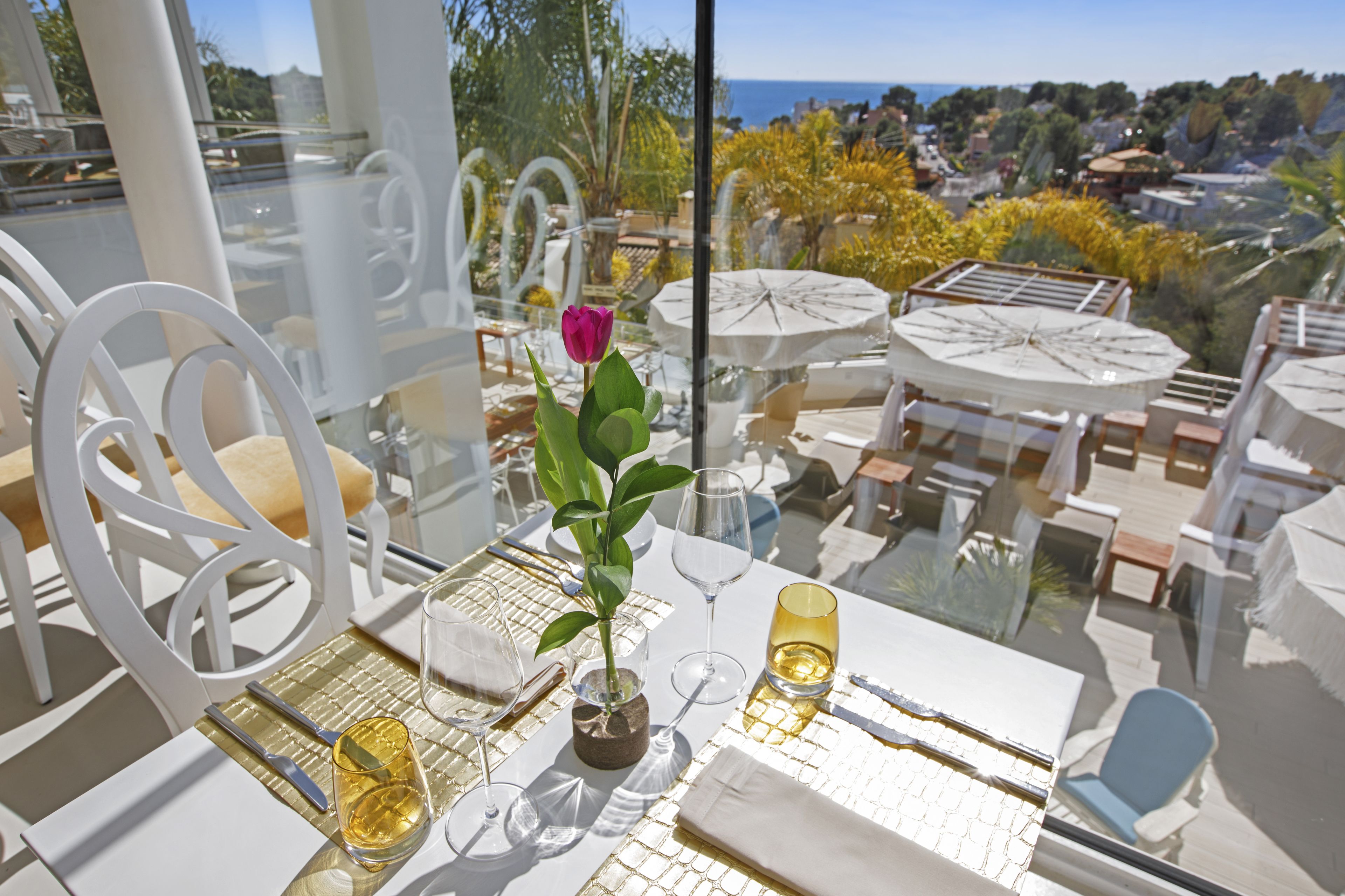 Un área de comedor al aire libre con sillas y mesas blancas, una rosa en un jarrón, y vistas al mar y las colinas.