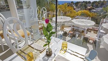 Un área de comedor al aire libre con sillas y mesas blancas, una rosa en un jarrón, y vistas al mar y las colinas.