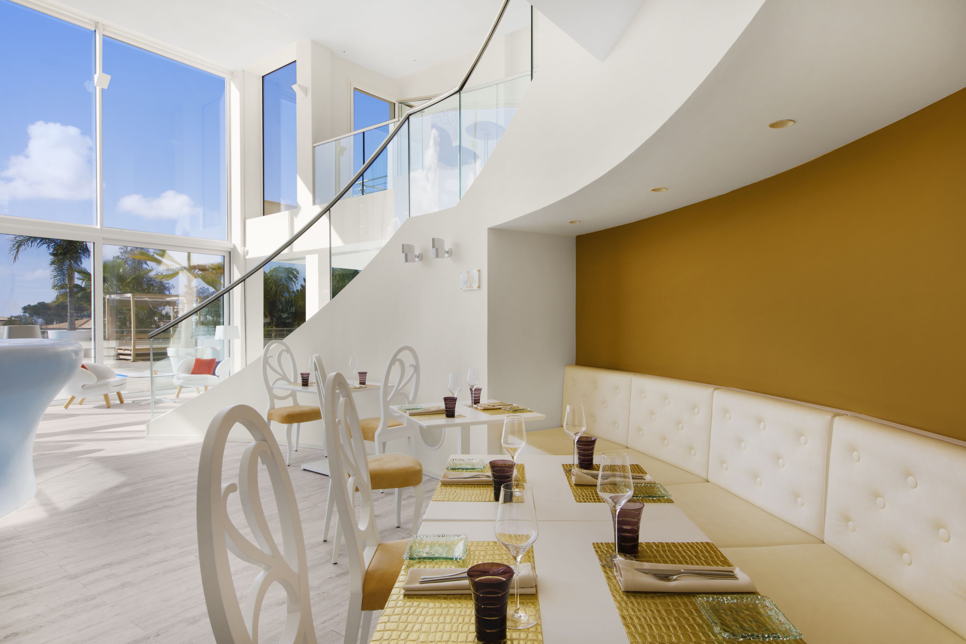 Un comedor moderno con mobiliario blanco, una escalera curva y grandes ventanales que ofrecen vistas al exterior.