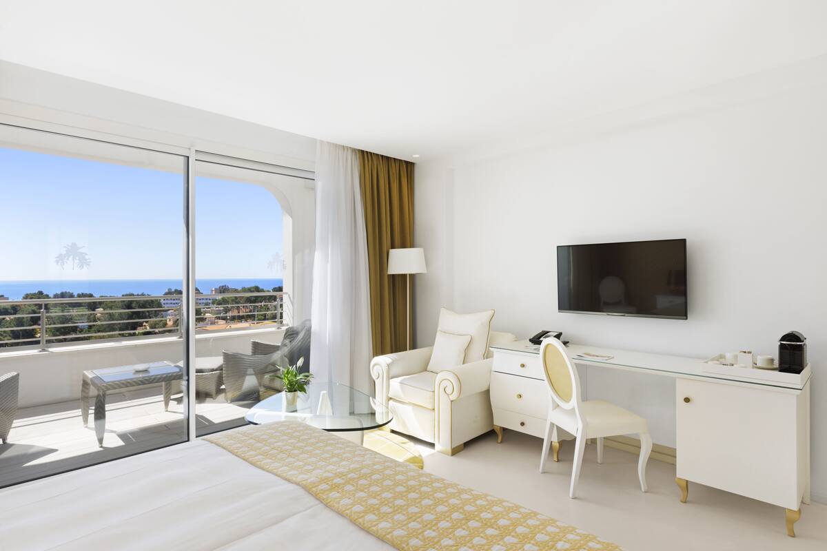 A modern hotel room with a large bed, a desk, a chair, a TV, and a balcony with a view of the ocean.