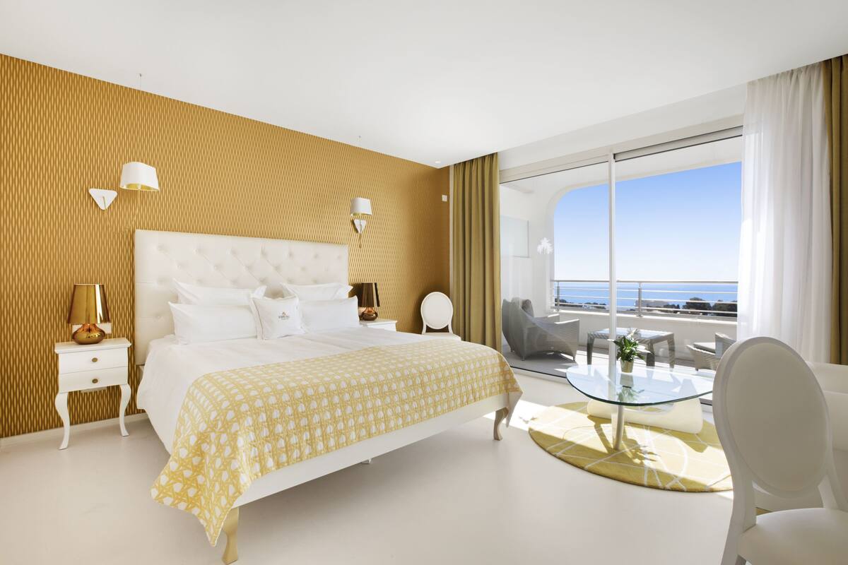 A modern hotel room with a large bed, a glass table, and a view of the ocean.
