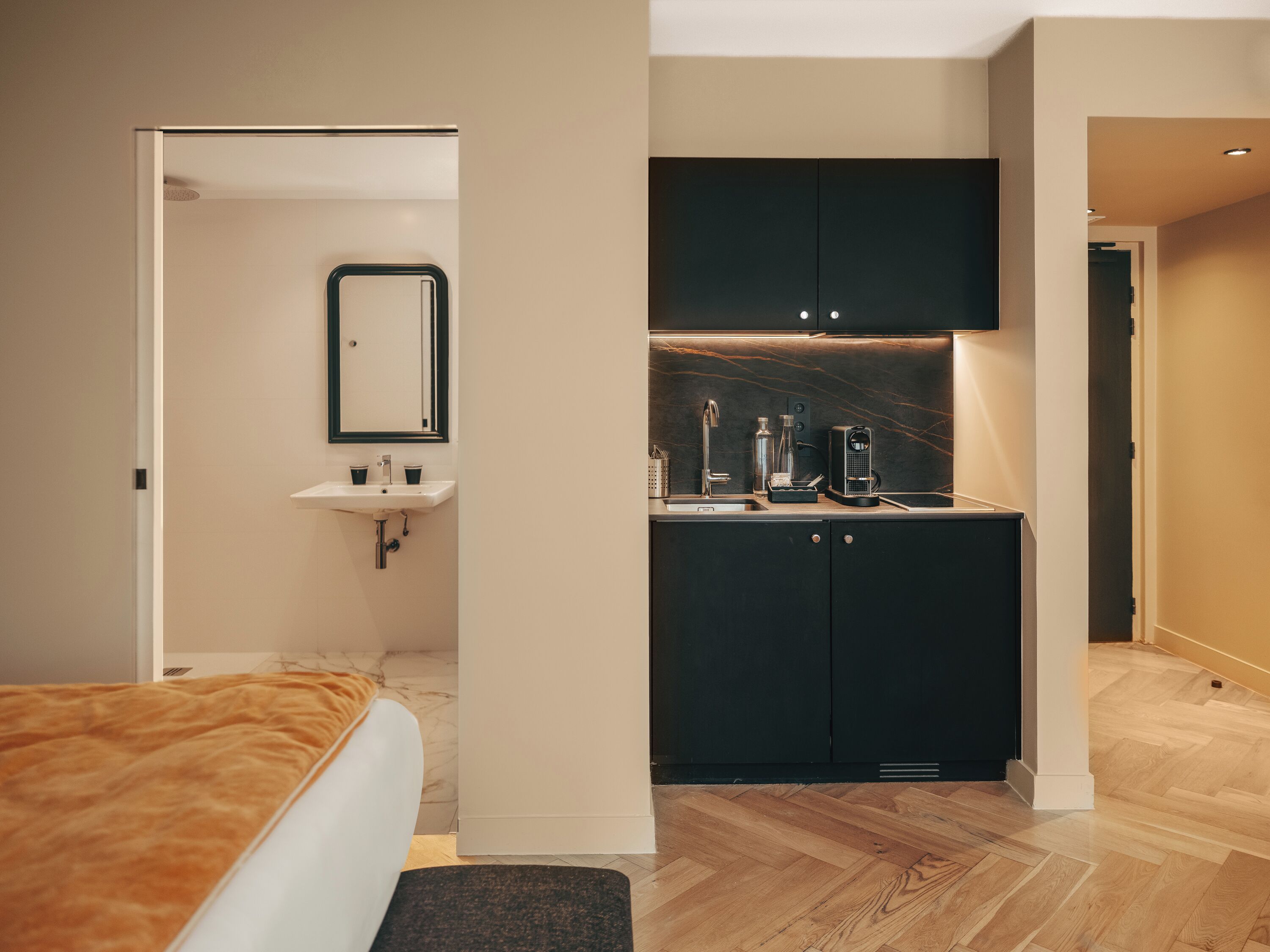 A modern hotel room with a bed, a bathroom with a sink and mirror, and a kitchenette area.