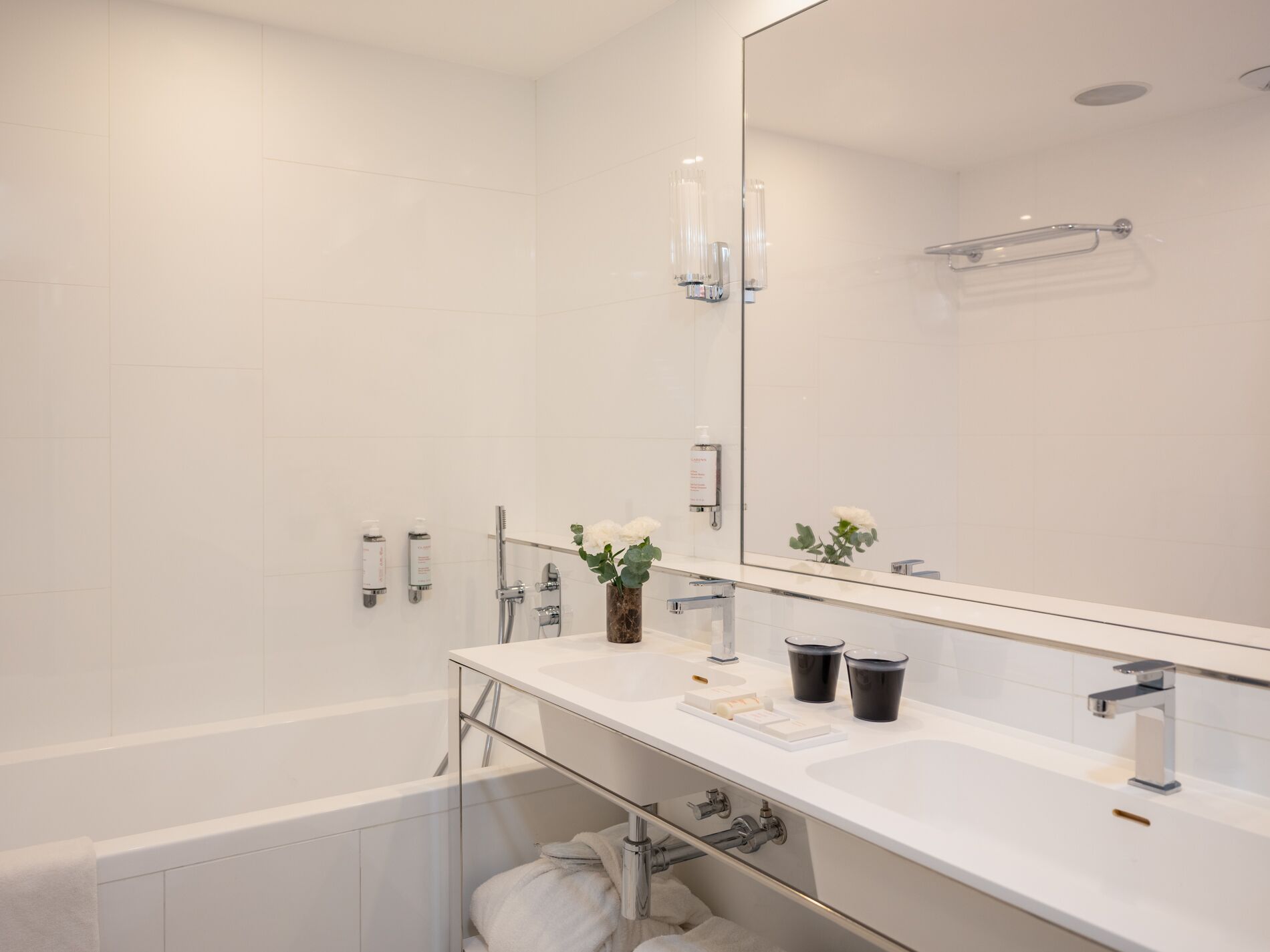 Un bagno moderno con un ampio specchio, piastrelle bianche, cabina doccia in vetro e una vasca da bagno bianca.