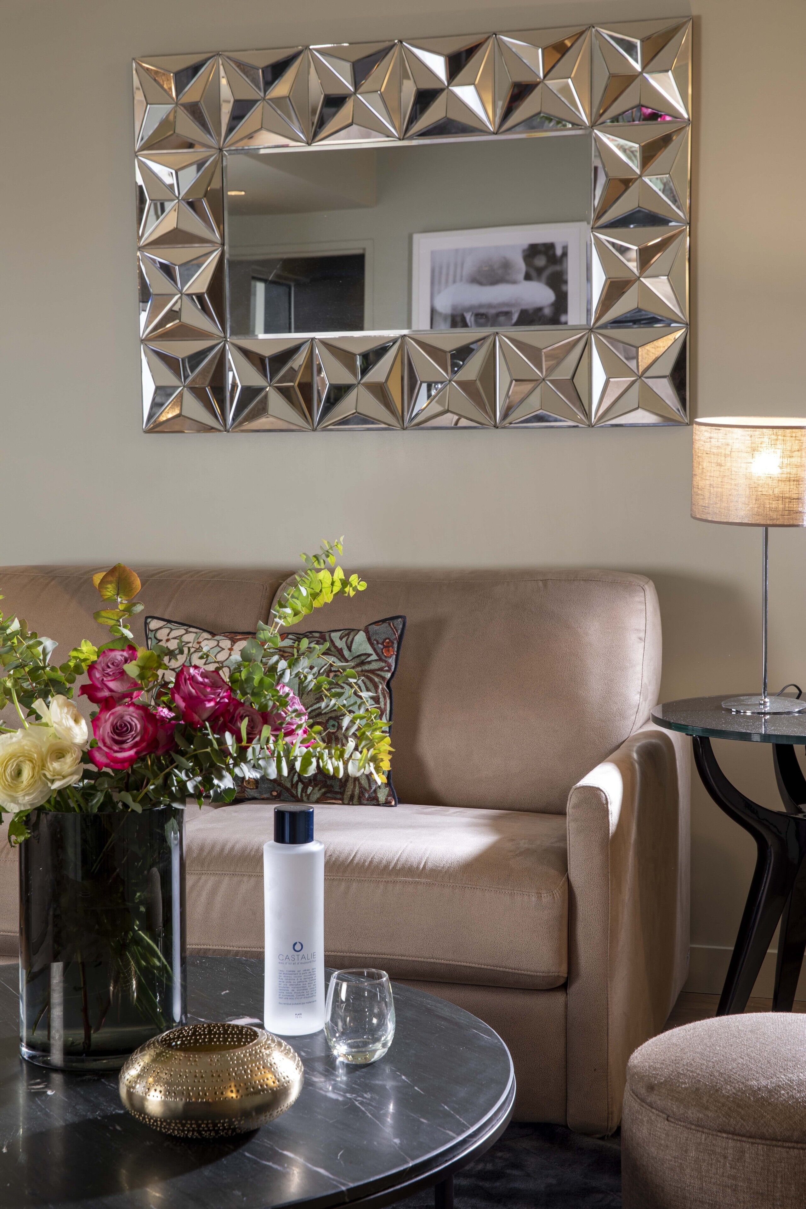 Ett vardagsrum med en beige soffa, ett runt soffbord med en vas med blommor, en dekorativ spegel och en lampa.