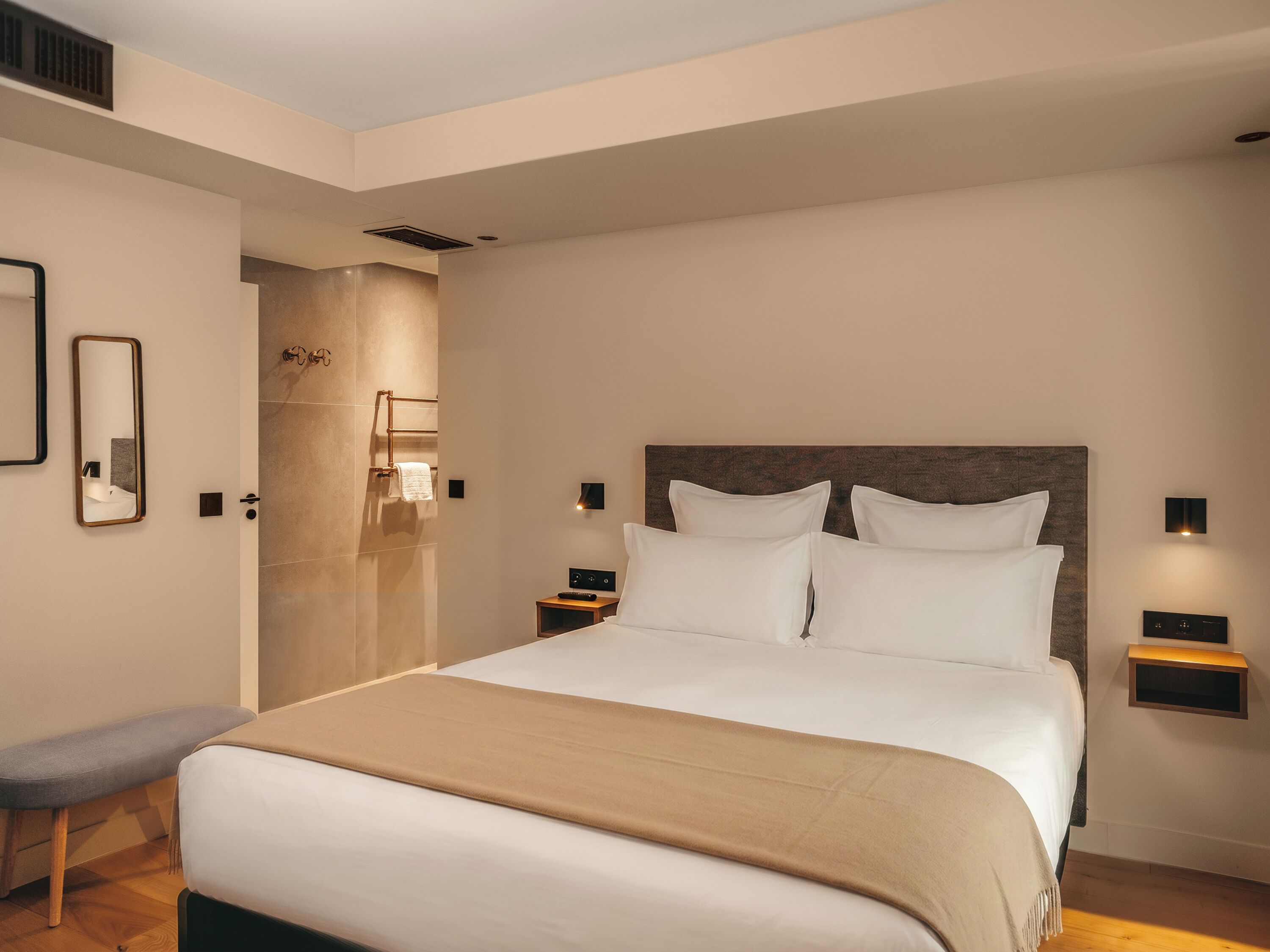 Quarto de hotel moderno com uma cama grande, mesas de cabeceira, um banco e área de chuveiro.