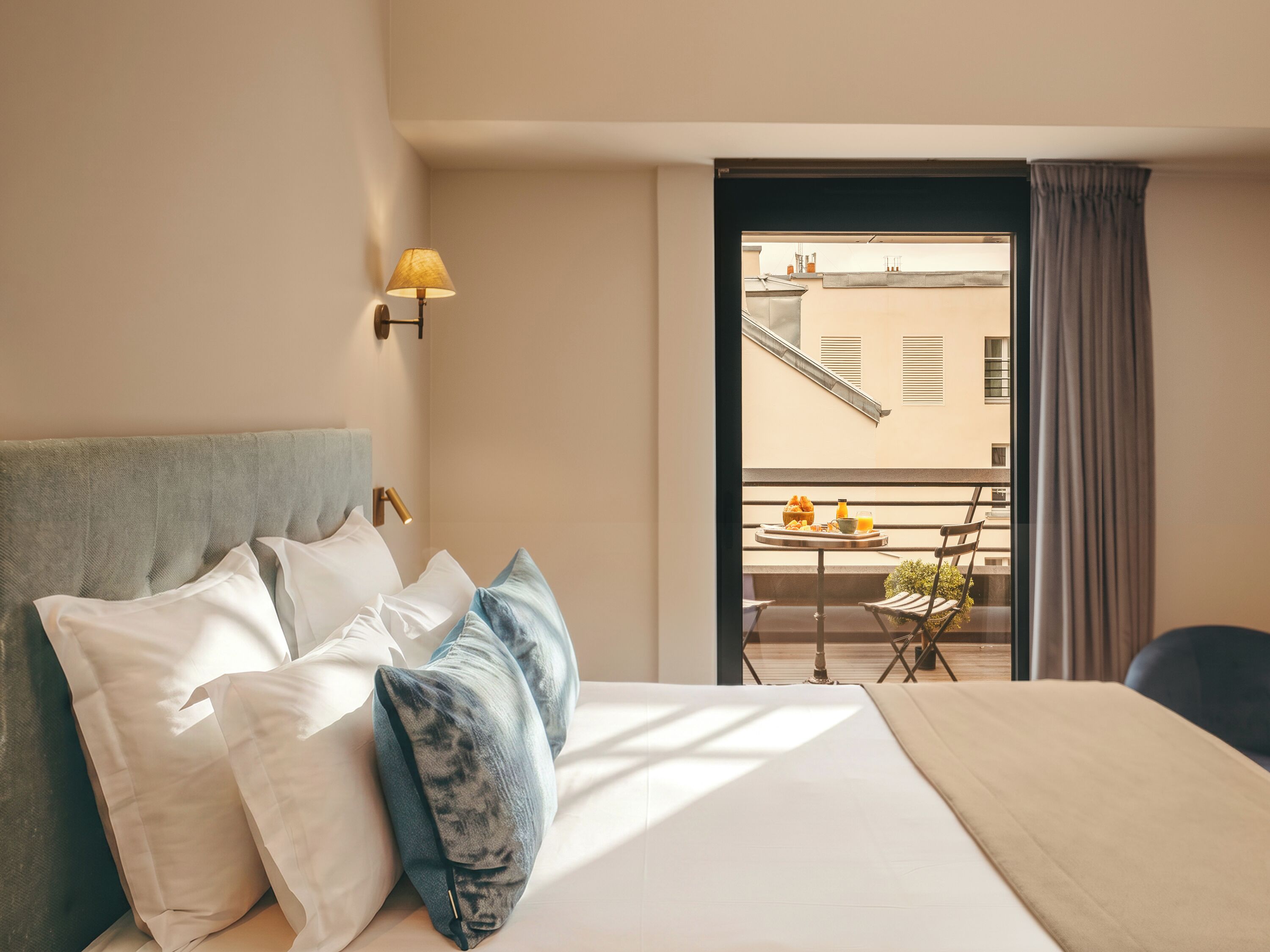 Camera d'albergo con letto, cuscini, comodino, lampada, balcone con tavolo e sedie, e vista sugli edifici.