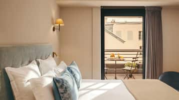 Camera d'albergo con letto, cuscini, comodino, lampada, balcone con tavolo e sedie, e vista sugli edifici.