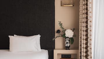 A neatly made bed with white linens, a dark textured headboard, a bedside table with a vase of white flowers, and a book titled "LOUIS VUITTON CATWALK" on the shelf.