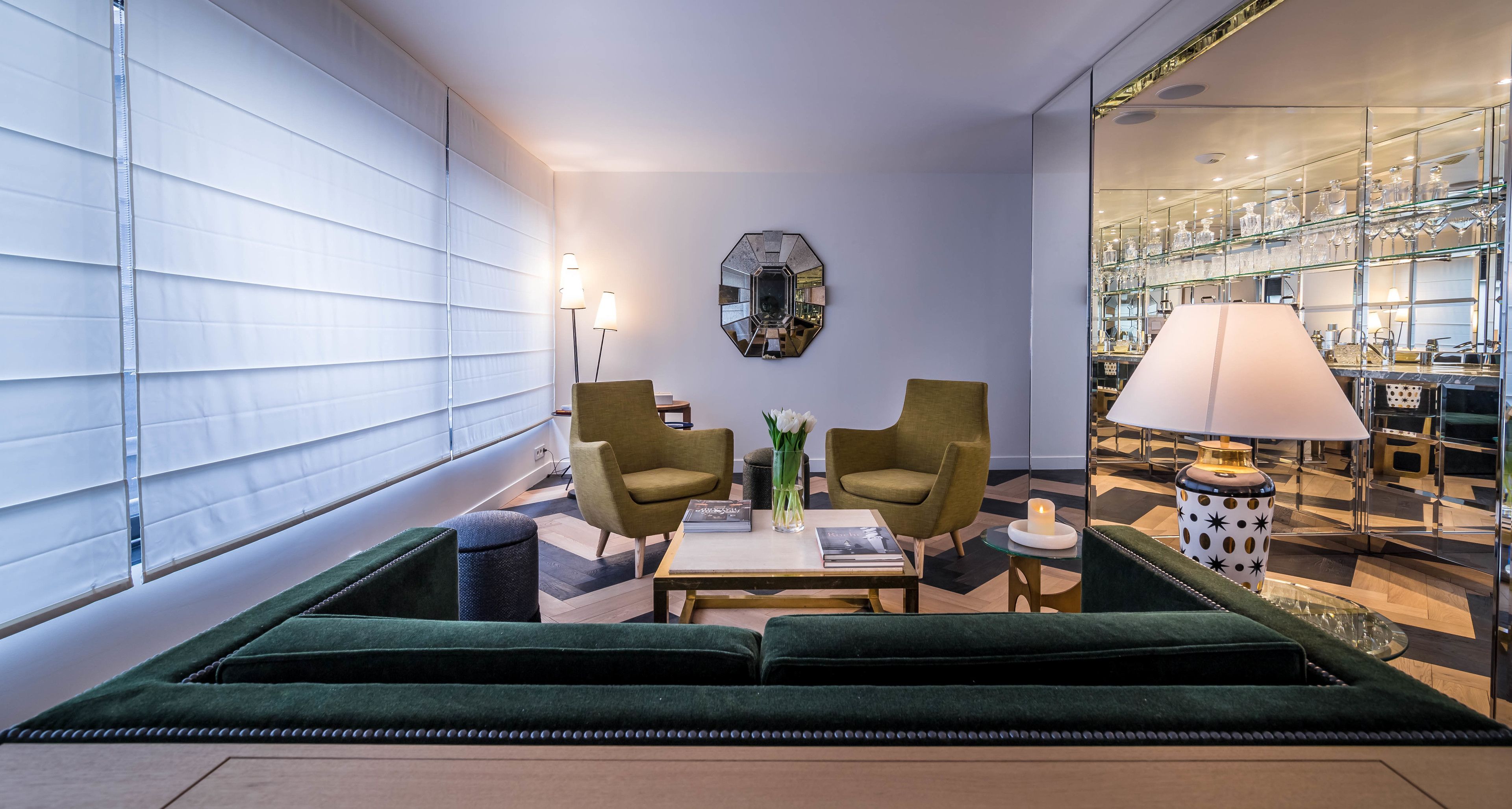 A modern living room with a green sofa, two armchairs, a coffee table, and a side table with a lamp. A mirror and a glass display cabinet are also visible.
