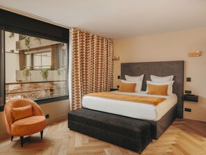 Ένα σύγχρονο δωμάτιο ξενοδοχείου με ένα μεγάλο κρεβάτι, έναν πάγκο, μια καρέκλα και θέα σε ένα μπαλκόνι με φυτά.