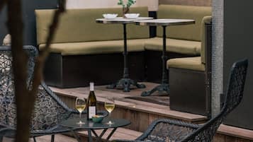 An outdoor seating area with wicker chairs, a wooden table, and a small table with a bottle of wine and glasses.