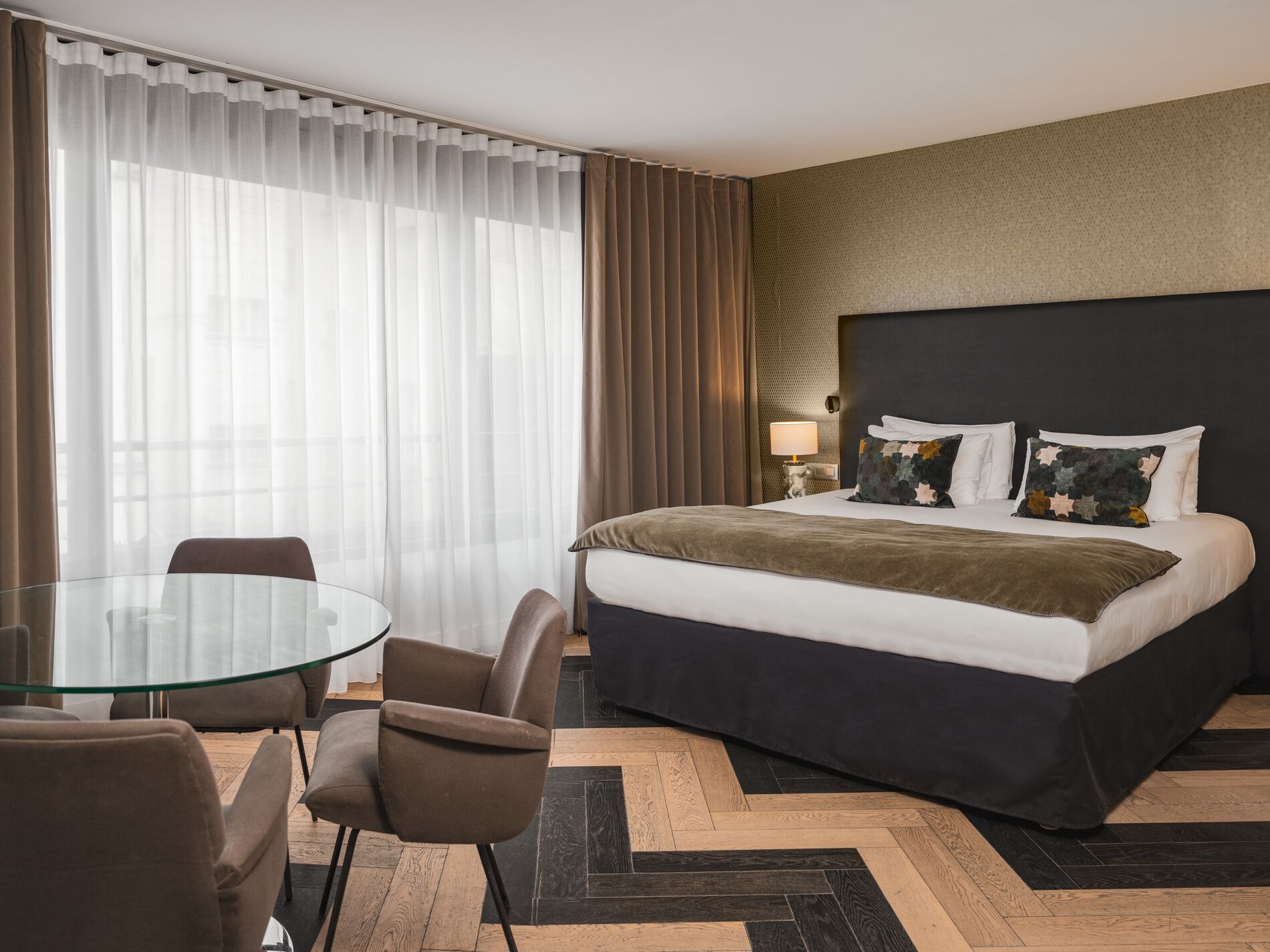 A hotel room with a bed, a round glass table, two chairs, a lamp, and a view of the outdoors through sheer curtains.