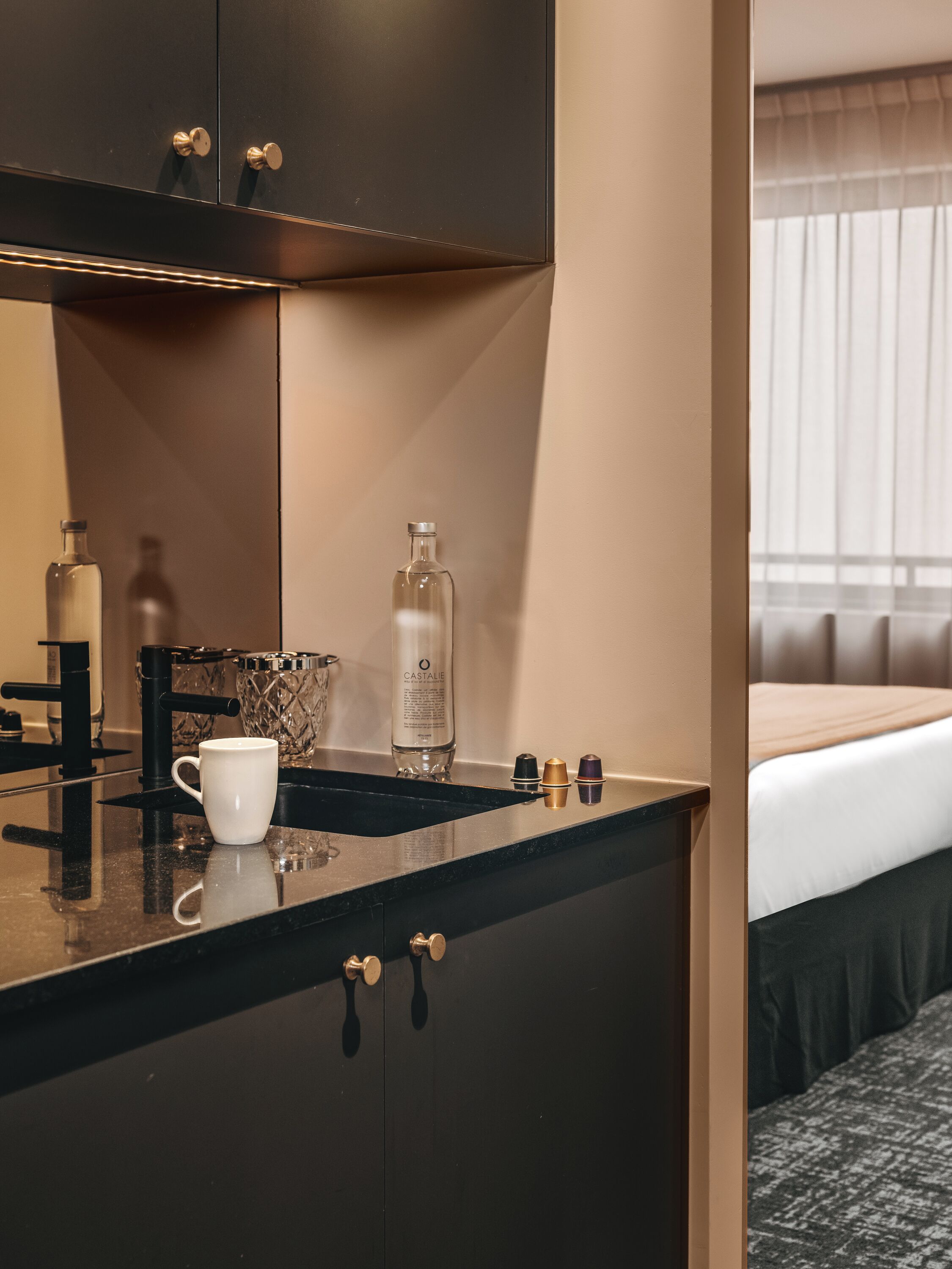 A modern hotel room with a dark-colored minibar, a white coffee cup, a bottle of Castalie, and a bed with a white comforter.