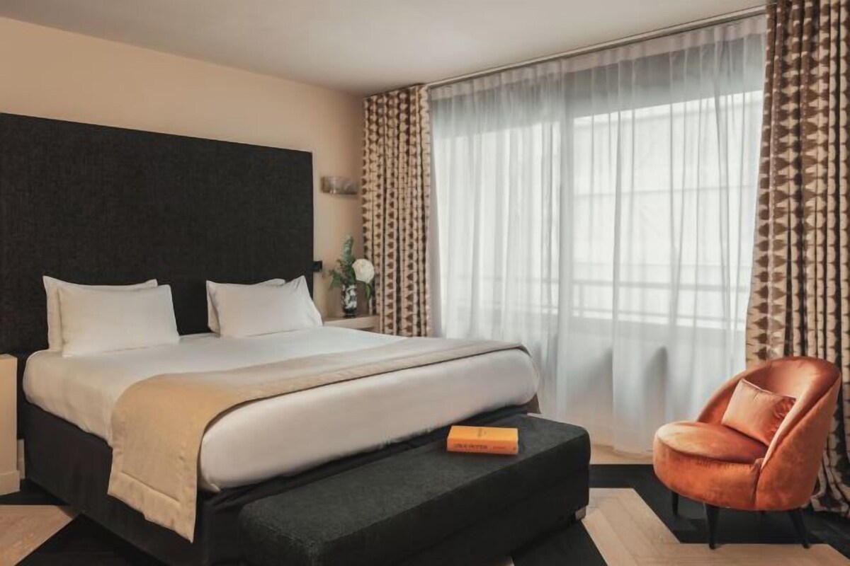 A modern hotel room with a large bed, a side table, a chair, and a large window with sheer curtains.