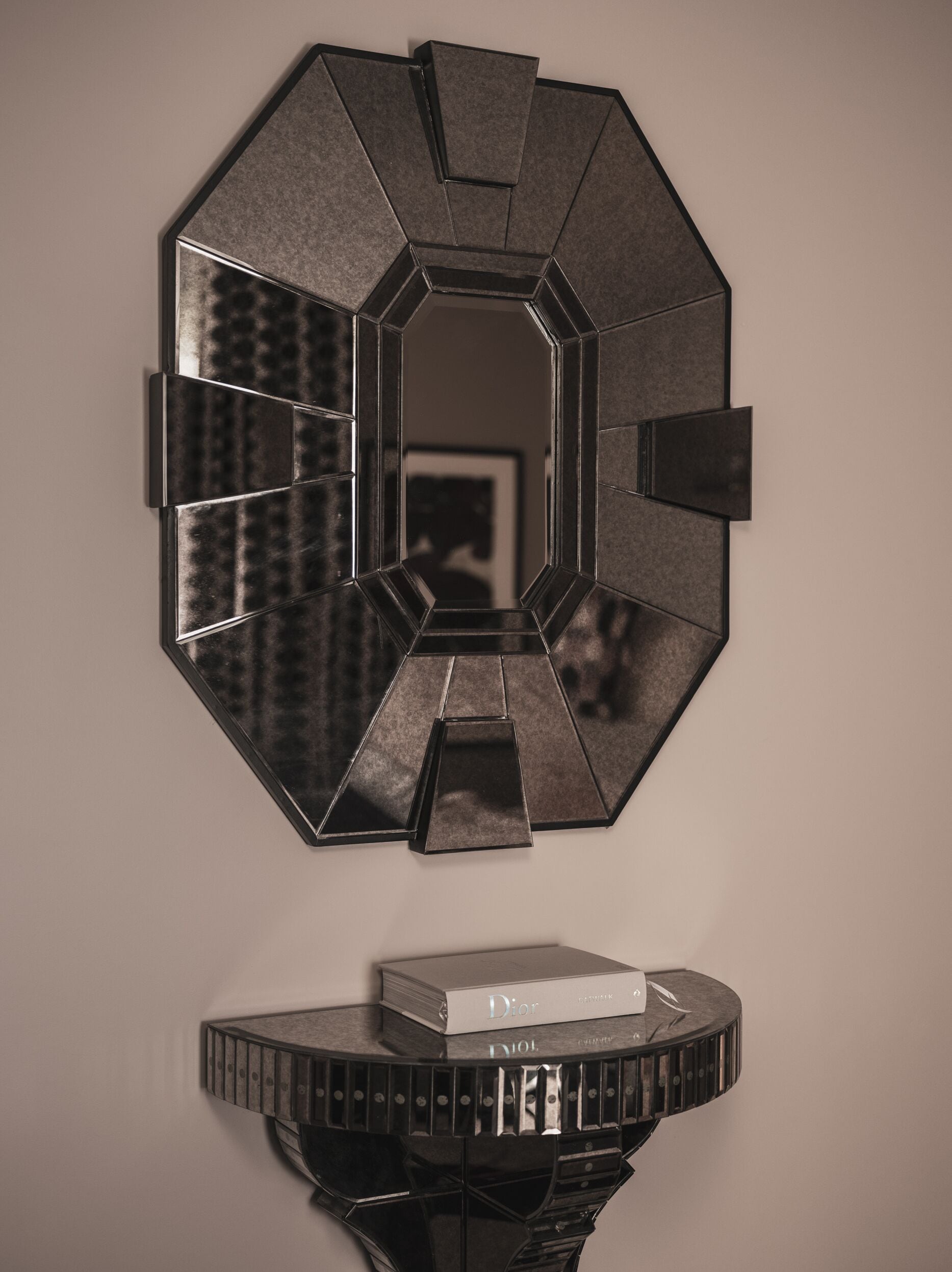 Une œuvre d’art géométrique pour mur, composée d’un miroir central encadré d’un cadre hexagonal. En dessous, une console noire supporte un livre et une paire de ciseaux.